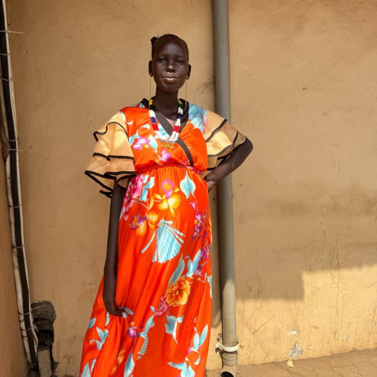 A person in a vibrant orange floral dress posing outdoors against a beige wall.